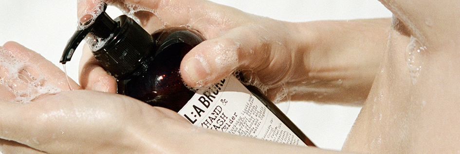 Eine Person hält unter fließendem Wasser eine braune Pumpflasche von L:A Bruket in den Händen, während Seifenschaum die Haut bedeckt.
