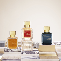 Drei Parfümflakons von Maison Francis Kurkdjian Paris stehen nebeneinander auf einer stilisierten Oberfläche mit Marmor- und Goldmustern. Von links nach rechts: ‚Grand Soir‘ in Bernsteinfarbe, ‚À la rose‘ in zartem Rosa mit rotem Etikett und ‚Oud‘ in dunklem Blau. Alle Flakons haben einen goldenen Verschluss.