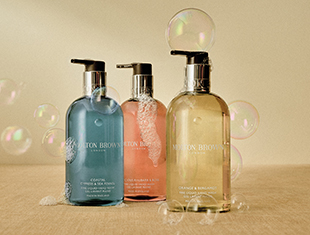 Drei Flüssigseifenspender von Molton Brown in Blau, Rosa und Honiggelb stehen nebeneinander auf einer beigen Oberfläche. Auf den durchsichtigen Flaschen bilden sich zarte Seifenblasen. Um die Produkte herum schweben weitere schillernde Blasen, was der Szene eine frische und saubere Ausstrahlung verleiht.