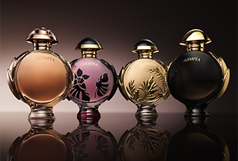 Vier elegante Parfumflakons der Linie 'Olympea' stehen nebeneinander auf reflektierendem Untergrund. Die Flakons sind rundlich mit goldenen oder bronzefarbenen Verschlüssen und tragen florale oder grafische Muster in verschiedenen Farbtönen: Beige, Violett, Gold und Schwarz.