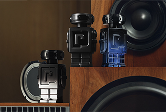 Drei schwarze Parfumflakons in Form von Roboterfiguren mit dem Buchstaben 'P' auf der Brust stehen zwischen großen Lautsprechern aus Holz. Die mittlere Flasche leuchtet leicht blau. Die Szene vermittelt einen modernen, technologischen Look mit Musikthema.