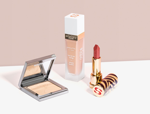 Drei dekorative Kosmetikprodukte von Sisley Paris auf heller Fläche vor rosa Hintergrund: ein geöffneter silberner Kompaktpuder mit Spiegel, ein Fläschchen flüssiges Make-up mit weißem Deckel und beiger Aufschrift, sowie ein geöffneter goldener Lippenstift mit rotem Farbton und gemustertem Deckel mit rotem 'S'-Logo.