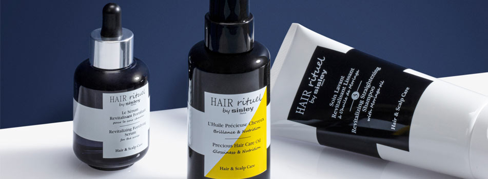 Drei Haarpflegeprodukte aus der Linie 'Hair Rituel by Sisley' stehen auf einer weißen Fläche vor einem dunkelblauen Hintergrund. Links eine dunkle Pipettenflasche mit Serum, in der Mitte eine Sprühflasche mit gelbem Etikett für ein Pflegeöl, rechts eine weiße Tube mit schwarzem Etikett. Die Produkte sind für Haar- und Kopfhautpflege konzipiert und tragen Beschriftungen in Englisch und Französisch.