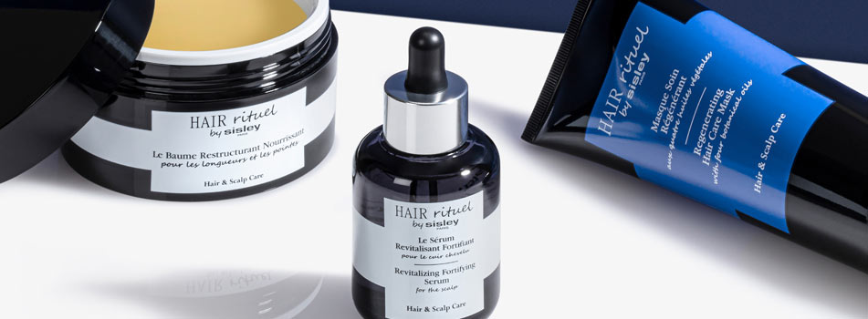 Drei Haarpflegeprodukte aus der Linie 'Hair Rituel by Sisley' auf einer weißen Fläche vor blauem Hintergrund. Links ein geöffneter Tiegel mit gelblicher Creme, in der Mitte eine Pipettenflasche mit Serum, rechts eine blaue Tube mit schwarzem Verschluss. Die Etiketten tragen Produktnamen auf Französisch und Englisch, darunter ein revitalisierendes Serum, eine nährende Haarmaske und eine restrukturierende Pflege.