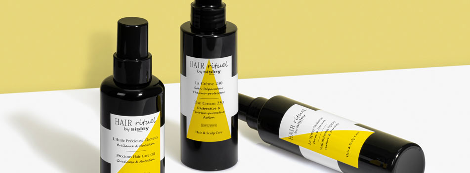 Drei schwarze Flaschen aus der Haarpflegelinie 'Hair Rituel by Sisley' mit weißen Etiketten und gelben Akzenten stehen bzw. liegen auf einer weißen Fläche vor einem gelben Hintergrund. Zu sehen sind unter anderem ein Precious Hair Care Oil, eine Flasche mit der Aufschrift 'La Crème 230' sowie ein weiteres Pflegeprodukt. Die Anordnung ist modern und minimalistisch.