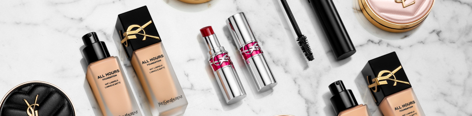 Make-up-Produkte von Yves Saint Laurent auf einer weißen Marmoroberfläche, darunter zwei Flaschen flüssige Foundation, zwei silberne Lippenstifte, eine Mascara und ein schwarzes Puderdöschen mit goldenem YSL-Logo.